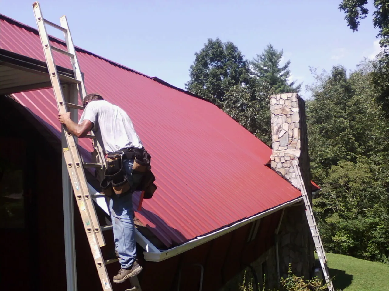 Roofer on a ladder beside a red metal roof and stone chimney — Soffit & Fascia Repair by Roofing Brothers in Gaines